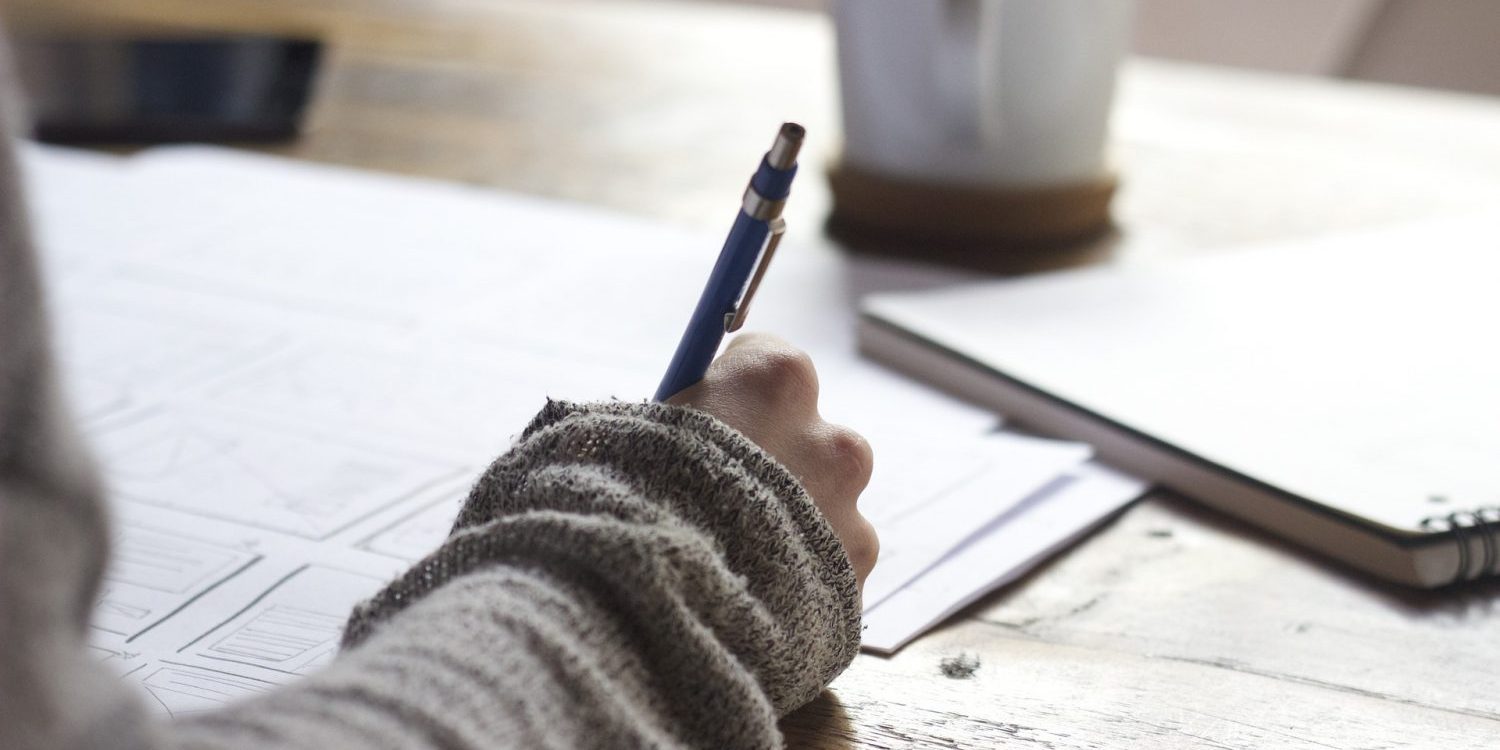 Woman's hand writing on paperwork at table with cup of coffee and notebook.